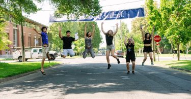 A group of Whitman students photographed mid-jump and smiling beneath a Whitman College banner stretched across a main campus street.