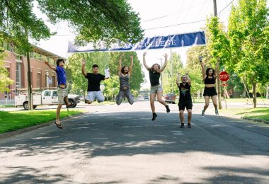 A group of Whitman students photographed mid-jump and smiling beneath a Whitman College banner stretched across a main campus street.