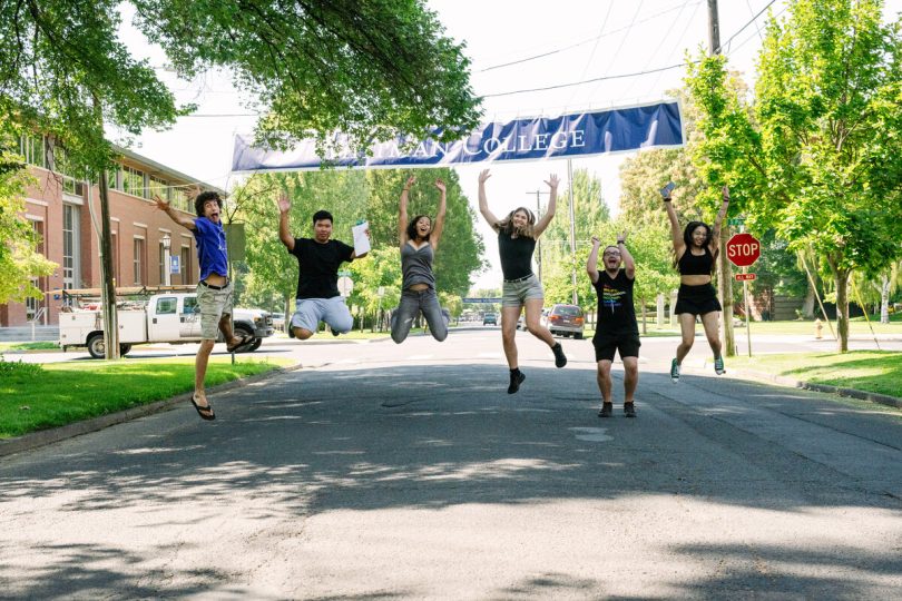 A group of Whitman students photographed mid-jump and smiling beneath a Whitman College banner stretched across a main campus street.