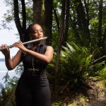 Student plays the flute in the woods with a lake to her back.