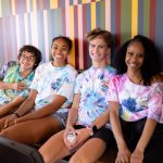 Students smiling at the camera, sitting on a bench in front of a brick wall.