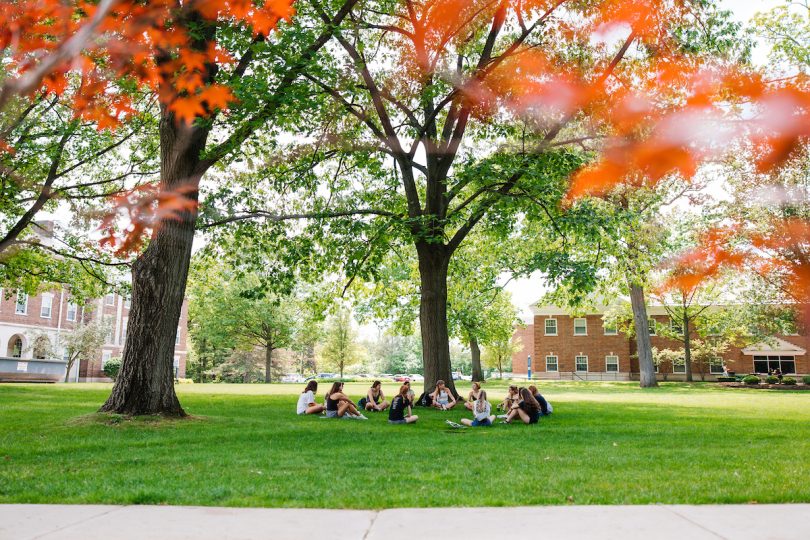 Ten students sit in a circle on a green lawn, surrounded by leafy trees and red brick buildings.