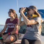 Two individuals in University of Redlands shirts are sitting on a boat looking through binoculars.